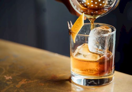 A glass of whiskey on the rocks with a large ice cube, orange peel garnish, and a decorative sip of liquor being poured from a decanter, ending with a flourish.