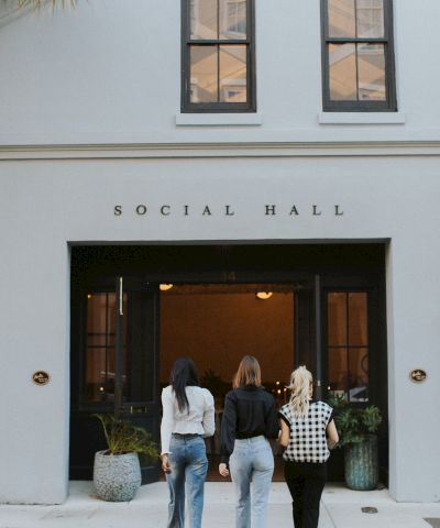 Three people walk into a light blue social hall with large glass doors, plant pots, and &ldquo;SOCIAL HALL&rdquo; above the entrance.