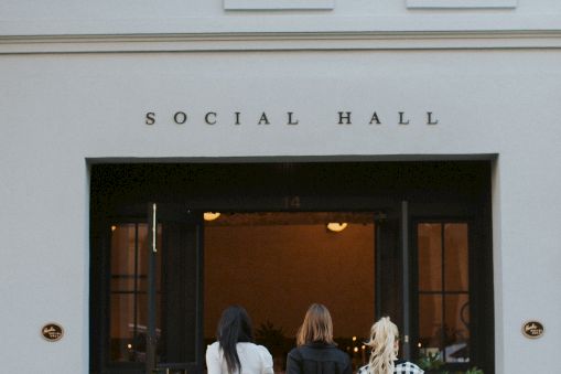 Three people walk into a light blue social hall with large glass doors, plant pots, and &ldquo;SOCIAL HALL&rdquo; above the entrance.