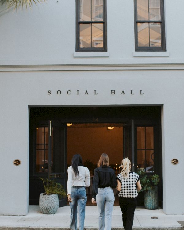 Three people walk into a light blue social hall with large glass doors, plant pots, and &ldquo;SOCIAL HALL&rdquo; above the entrance.