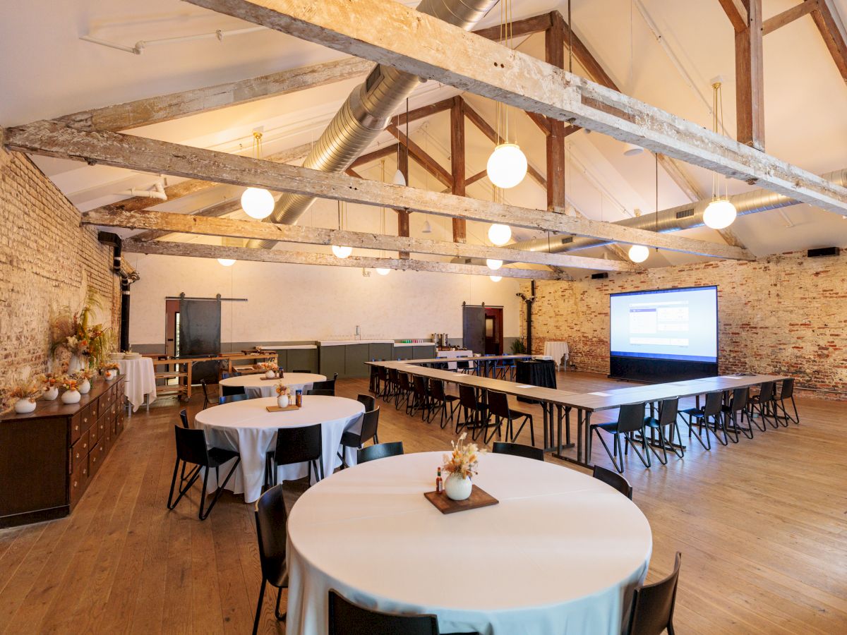 Wide rustic event space with round tables, white tablecloths, wooden chairs, exposed brick walls, and a projector screen at the far end.