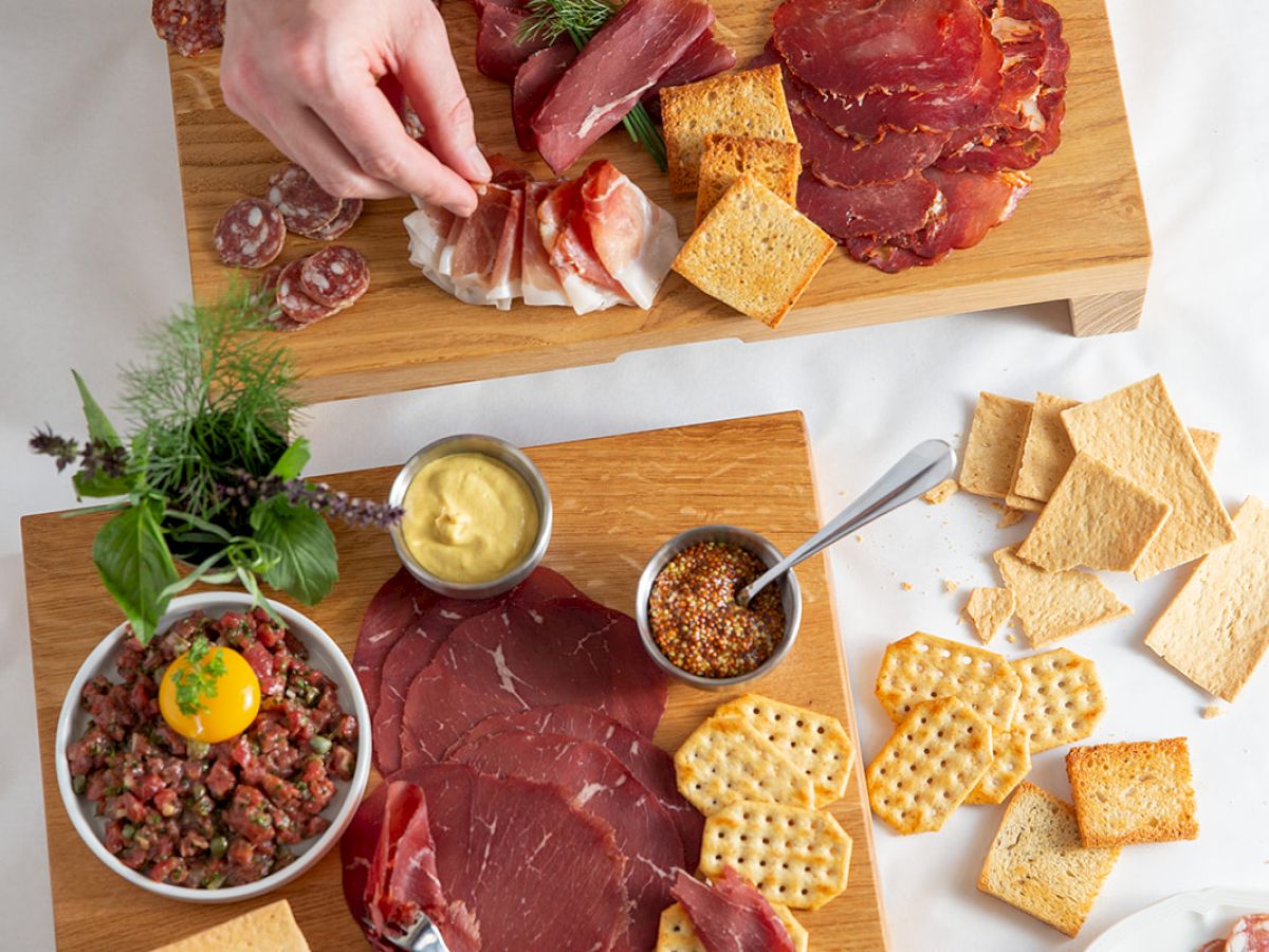 A wooden charcuterie spread with assorted meats, cheeses, crackers, dips, and a small salad, with hands arranging bites on boards.