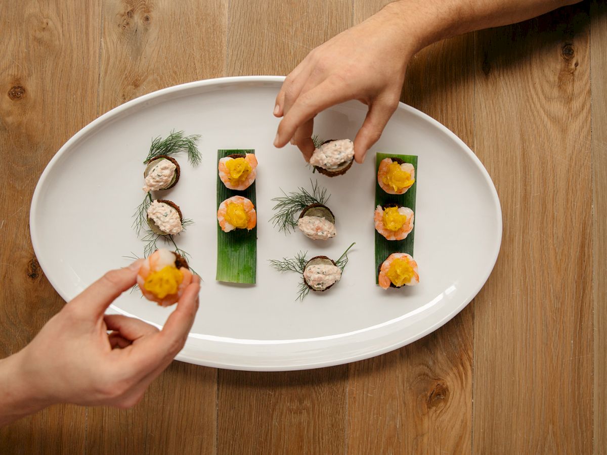 A white plate on a wooden table with neatly arranged bite-sized sushi pieces, hands assembling and garnishing with greens and citrus slices.