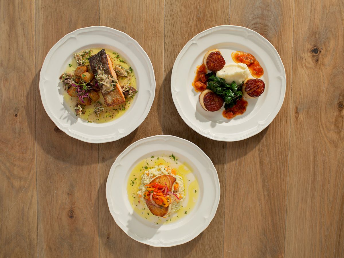 Three plated dishes on a wooden table: a ravioli-like pasta with greens, a dessert with berries and cream, and a sunny-side-up egg dish with shrimp, all neatly arranged.