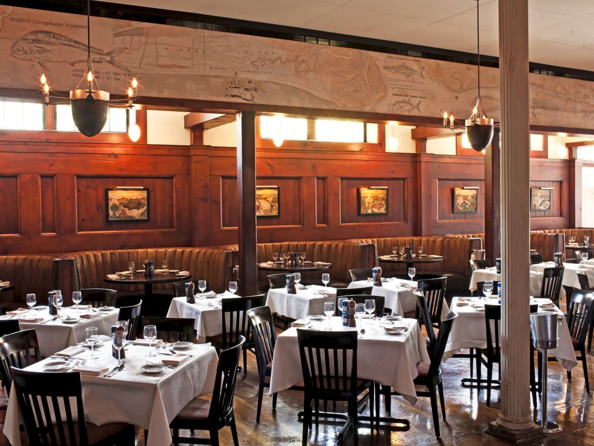 A stylish, upscale dining room with white-tablecloths, dark wooden chairs, booths along walls, soft lighting, and framed art on wood-paneled walls.