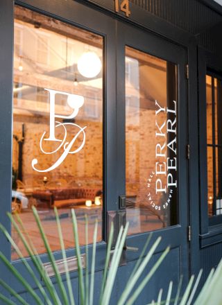 A stylish storefront with large glass doors, warm interior lighting, and white lettering on the window; plants frame the entrance.