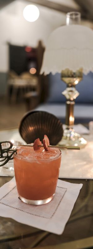 A pink cocktail with a pineapple leaf garnish sits on a napkin at a rounded table, a seashell decor piece and lamp in the blurred background, and a cozy restaurant vibe.