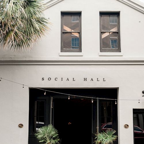 A light-gray building labeled &ldquo;Social Hall&rdquo; with string lights, two potted palms, and an open doorway, palm tree on left.