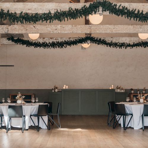 A decorated event hall with hanging garlands, round tables set with white tablecloths and chairs, ready for a formal gathering.