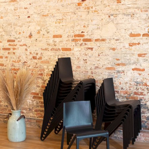 A neat stack of black chairs leaning against a rustic brick wall, with a lone chair in front and a vase of dried reeds nearby, in a minimal room.