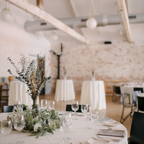 A formal dining setup with a round table, white linens, glassware, and a floral centerpiece in a bright, airy venue with brick walls.
