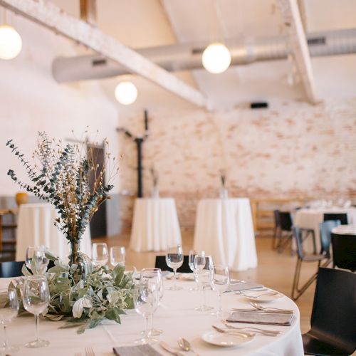 An elegant dining setup with a round white-tablecloth, white napkins, glassware, and floral centerpiece in a bright, modern banquet hall.