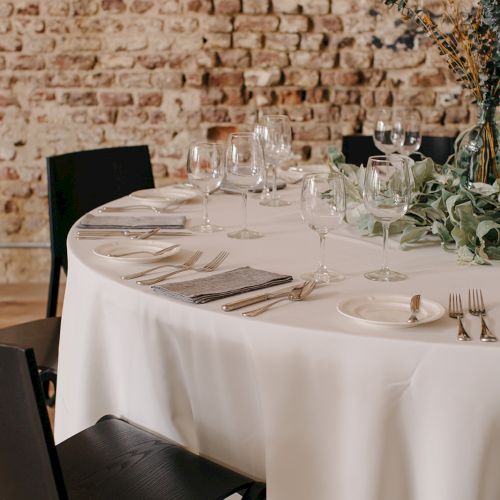 A neatly set dining table with white tablecloth, glassware, and place settings against a brick wall backdrop. Top it at 140 characters, always ending the sentence.A formal dining setup with a white tablecloth, multiple wine glasses, plates, cutlery, and a subtle centerpiece, ready for guests.