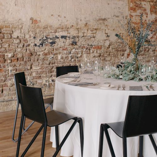 A round table with a white tablecloth sits against an exposed brick wall, surrounded by black modern chairs in a minimalist dining setup.