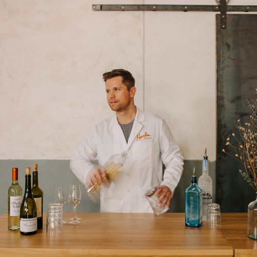 A man in a white lab coat mixes drinks behind a wooden bar with bottles, glassware, and dried flowers on the counter, smiling as he shakes.