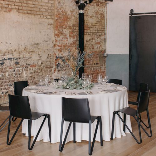 A round table with a white cloth, set for a formal dinner, surrounded by black chairs in a rustic brick-walled room with subdued d&eacute;cor.