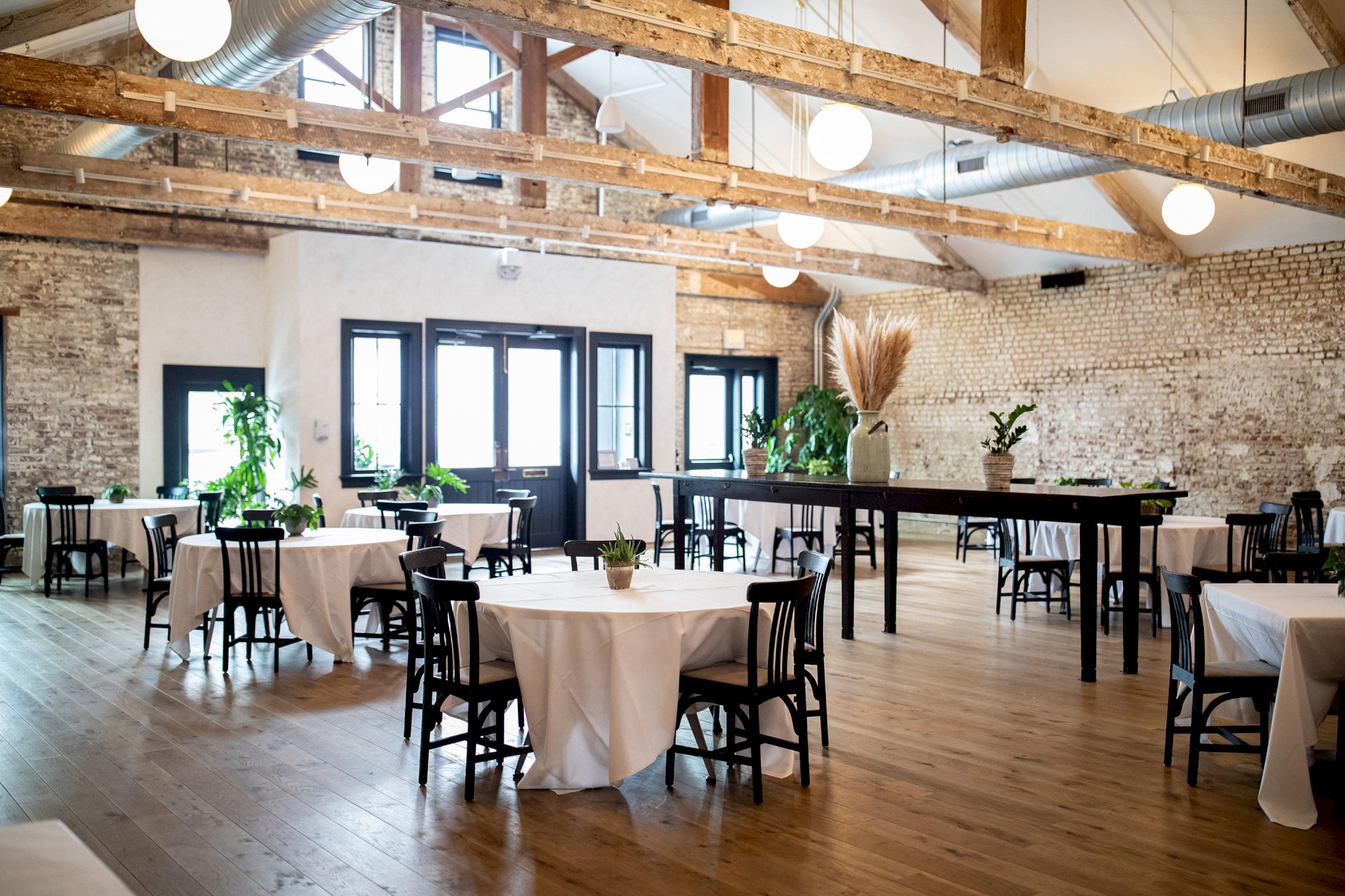 A bright, airy event space with exposed brick walls, wooden beams, and round tables covered in white cloths, set for a gathering.