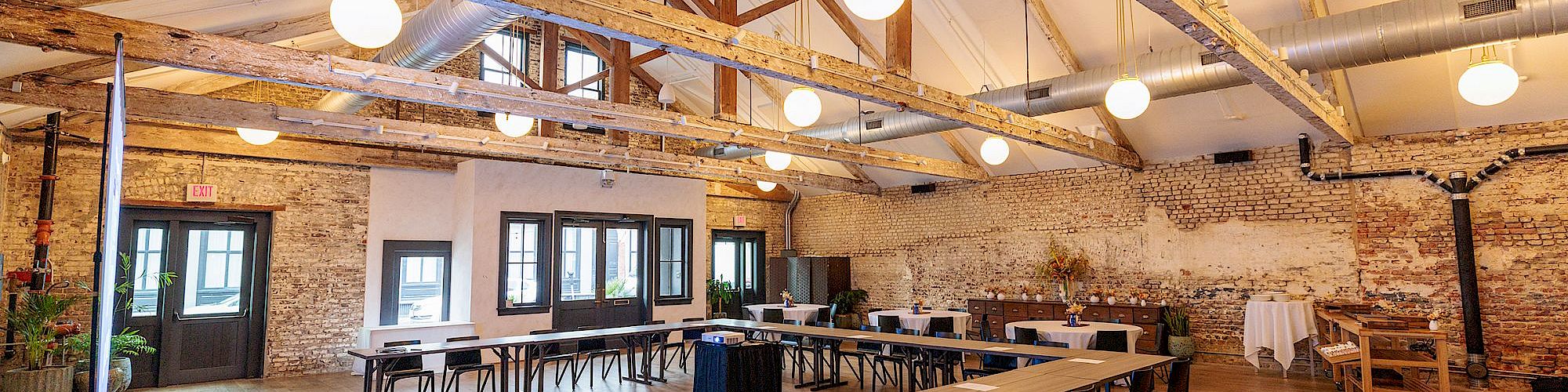 A spacious, well-lit workshop or meeting room with long tables, black chairs, exposed brick walls, wooden floors, and visible ceiling beams with round hanging lights. End the sentence.