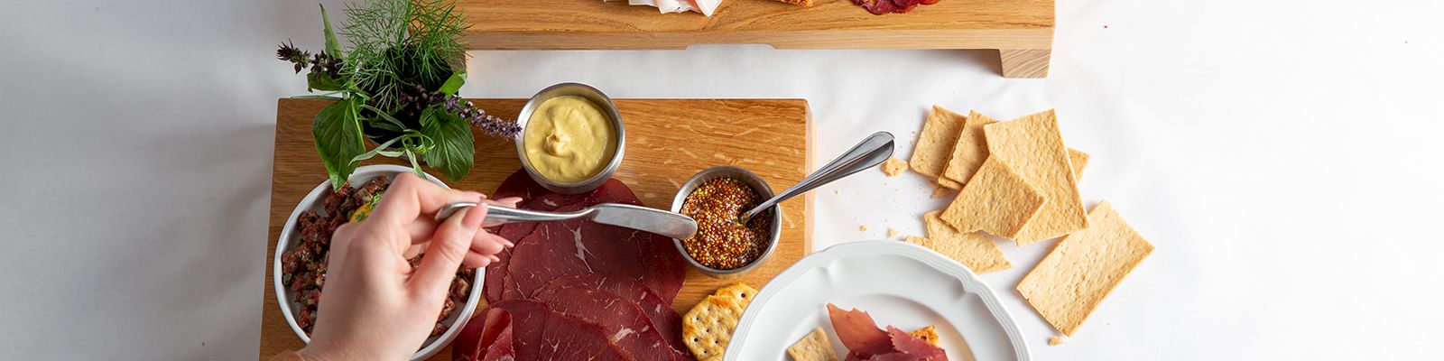 Hands assemble a charcuterie board with cured meats, bread, pickles/olives, and mustard dip on wooden boards.