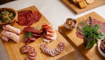 A charcuterie spread on wooden boards: sliced salami and cured meats, herbs, and a bowl of pepper, ready to eat.