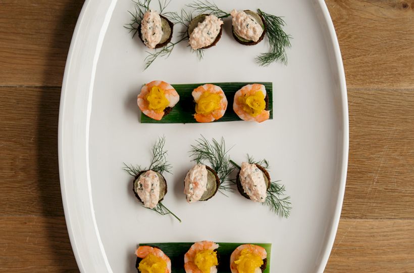 A white platter holding six bite-size appetizers: smoked salmon on avocado and shrimp with yellow garnish, garnished with dill.
