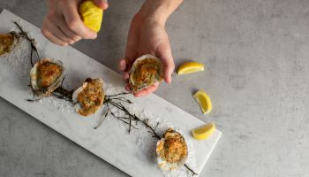 Hands hold baked oysters on a marble board, with lemon wedges and salt crystals on the tray.