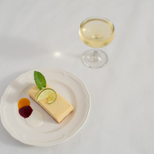 A glass of pale wine sits above a plate with a slice of cheese, lime wedge, mint leaf, and fruit.