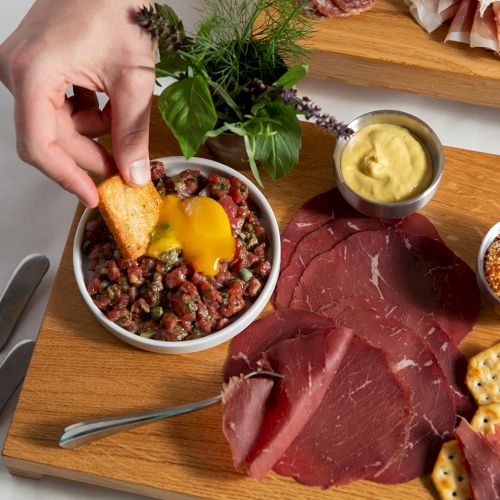 A charcuterie board with prosciutto, crackers, chopped meat, an egg yolk, and herb garnish on a wooden tray.