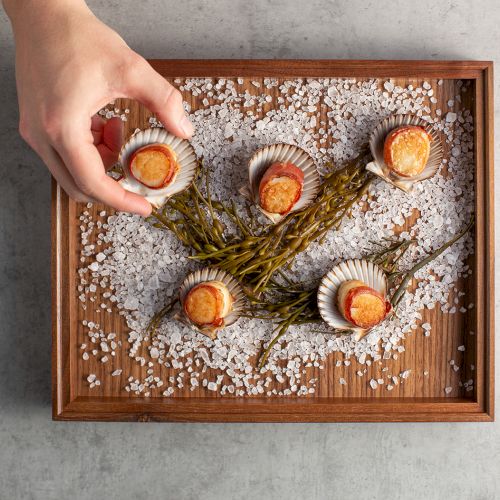 A hand arranges small golden bites in fluted shells on a wooden tray, surrounded by coarse salt and green stems.