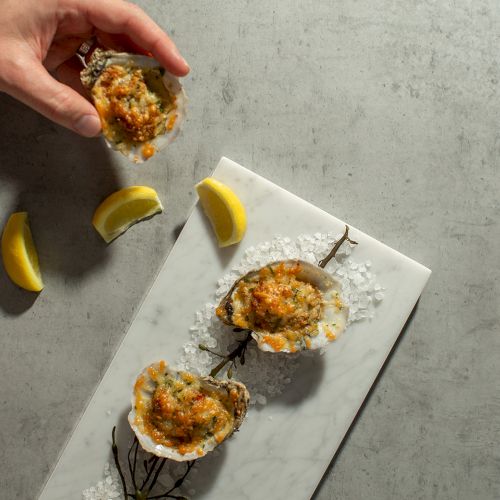 A hand sprinkles sauce/seasoning on oysters on ice with lemon slices on a marble board.