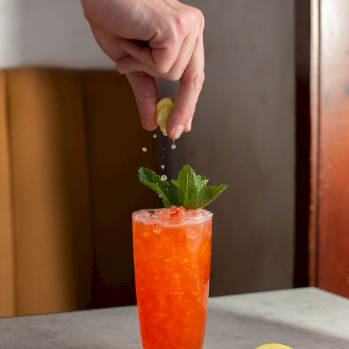 A hand squeezes lemon juice over a tall orange drink, garnished with mint, for a fresh cocktail.