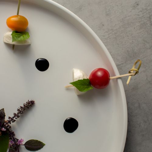 A white plate arranged with skewered cherry tomato and mozzarella, plus black olives and orange tomato bites around a floral garnish.