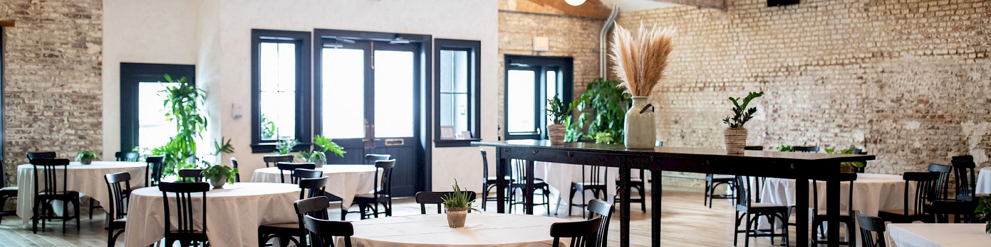 A bright industrial event hall with round tables set with white linens, black chairs, hanging lights, exposed brick, and plants.
